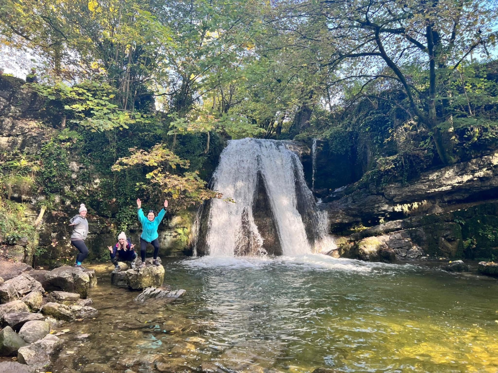 Three walkers with arms raised beside an autumn waterfall